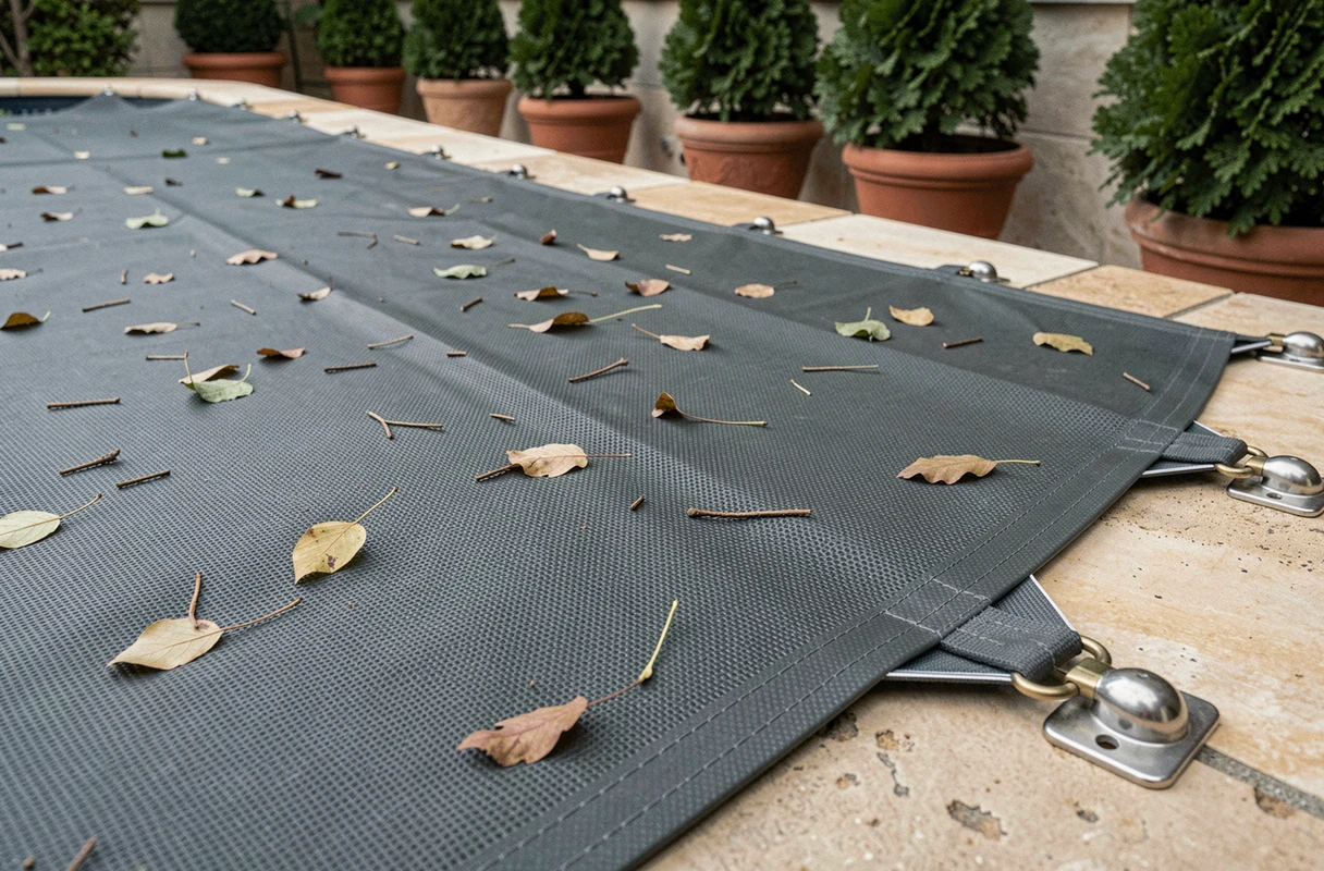 Gros plan sur une bâche de protection en PVC pour piscine avec feuilles mortes et fixations en laiton sur terrasse en pierre